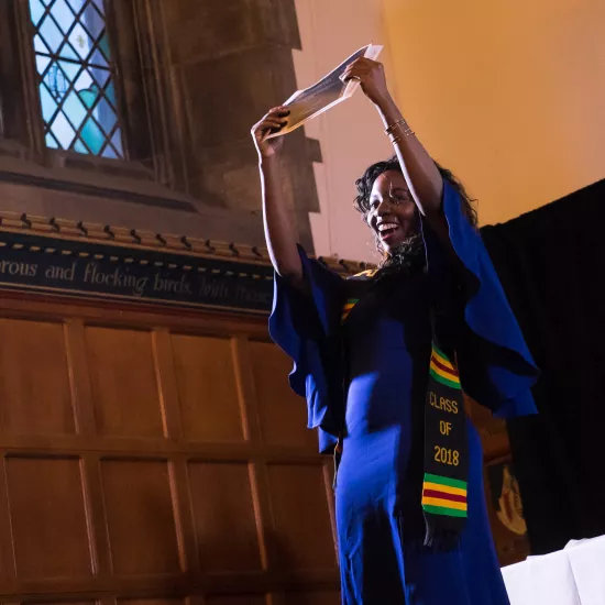 A Black woman wearing a convocation gown and holding up a diploma