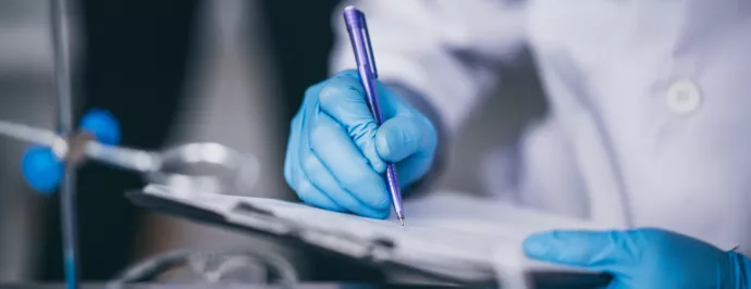 Closeup of a student in a white lab coat and gloves in a lab writing on a clipboard.