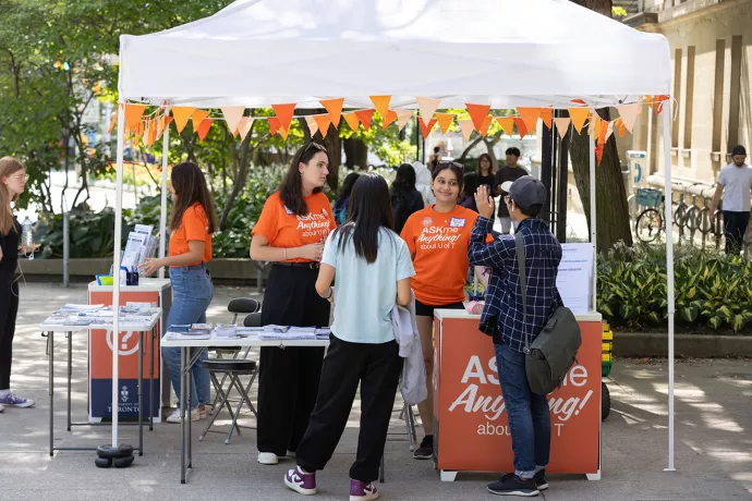 Students wearing orange Ask Me Anything t-shirts stand at an outdoor tent speaking with other students.