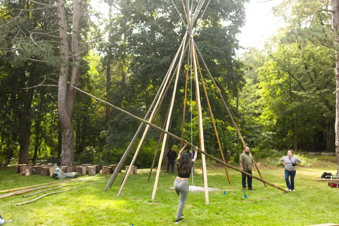 A woman holds a long wooden pole over her head as she walks it toward an unfinished Tipi structure