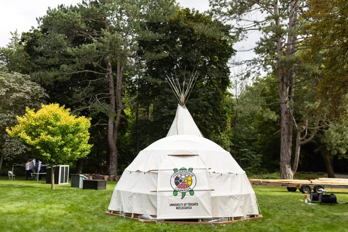 A photo showing a lowrise, domed teaching lodge made of white canvas and behind it can be seen the peak of the Tipi