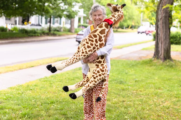 Anne Innis Dagg standing on a lawn wearing giraffe-print pants holding a large stuffed giraffe
