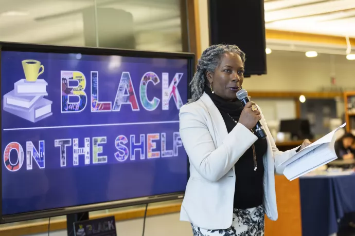 Heather Hines stands while holding a book and a microphone in front of a sign reading "Black on the Shelf"