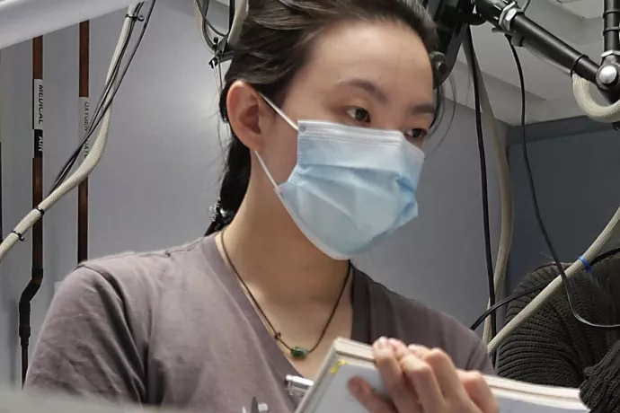 Student wearing blue surgical mask, jade necklace and brown t-shirt holding a notebook and pen, watching something off camera. Cables and unidentified equipment in the background.