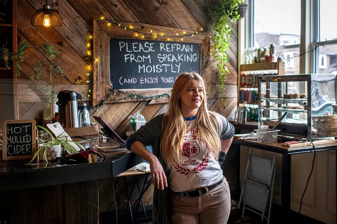 Sasha Steinberg standing by counter in restaurant, display of baked goods and four rows of shelves holding beverages behind her. Sign on counter says "Hot mulled cider" and chalkboard behind her on wall reads: Please refrain from speaking moistly (and enjoy your day).