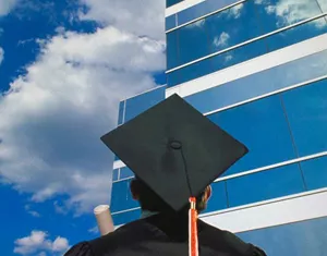 man in mortar board hat looking at tall building with blue sky in background