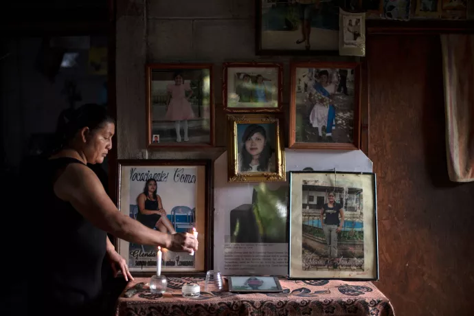 Woman standing at table, lighting candle, various pictures of daughter seen hanging on wall behind table.
