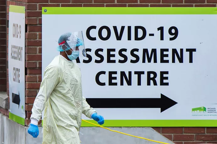 Person in protective gear walking by large sign that reads COVID-19 assessment centre