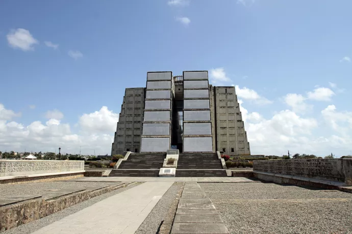 Image of large concrete structure in the distance, with long path leading to the stairs that lead to the structure.. Concrete structure has two stepped square columns in the front with concrete panels holding illegible inscriptions. The structure is split in the middle by a path.