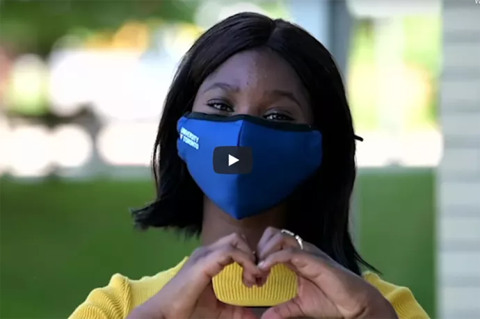 Girl wearing U of T face mask looking into camera, her hands making a heart symbol