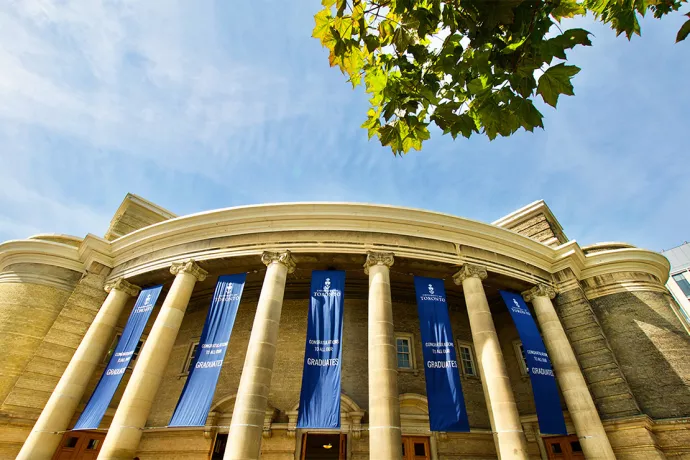 Image of Convocation Hall, looking up from the ground. Rounded entrance with tall concrete columns. Between each column is a blue banner hanging down reading Congratulations to all our graduates 
