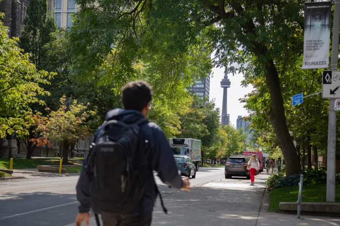 Man wearing backpack walking away from camera, down a tree-lined street with CN Tower in the distance
