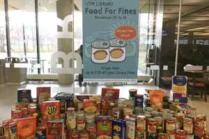 Stacks of canned food on a table with a sign that reads: UTM Library Food for Fines