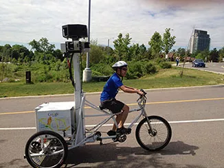 A smiling man pedals, pulling a large camera mounted to a post on a specialized bike used to capture images for Google Street View