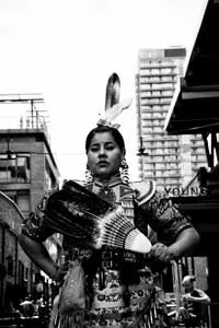 A woman wearing jingle dress regalia stands on a city street.