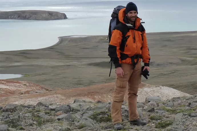 Igor Lehnerr wearing a red winter jacket with a black backpack standing on rocky, flat land with a lake in the background