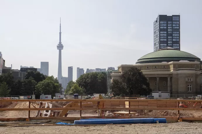 Construction site in foreground, with a pit dug, wooden fence in front of the pit with a digger in the pit. Background shows the CN tower and Convocation Hall
