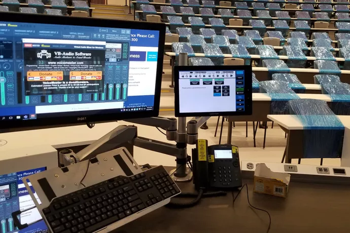 Two computer monitors on stand in front of classroom. Background shows lecture hall, with most chairs covered in blue plastic to ensure physical distancing.