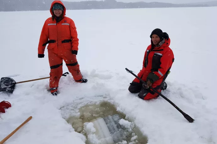 Two people in red winter coats and snow pants stand next to a square hole opening in the frozen lake.
