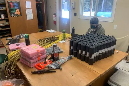 Person in toque and mask sitting at wooden table with cannisters, ropes and boxes on the table