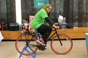 A woman wearing a tan hijab and a green shirt pedals a bike. The bike has a blender mounted over the back wheel blends a pink smoothie drink.