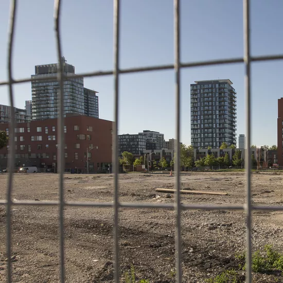 Photo looking through a wire fence at an empty lot with older low rise buildings and newer high rises in the background.