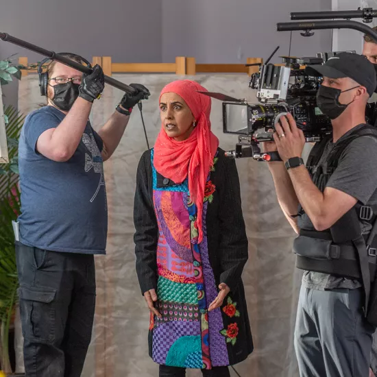 Zarqa Nawaz stands between a man holding a boom mic and two camera operators on a movie set.