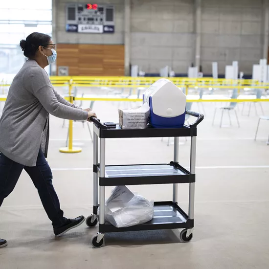 Woman pushes cart of medical supplied through RAWC gymnasium, which has rows of chairs physically distanced