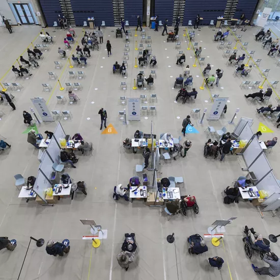 Overhead shot of chairs, physically distanced, in gymnasium, with people sitting in them after being vaccinated. Also seen are vaccination stations where people are receiving their dose.