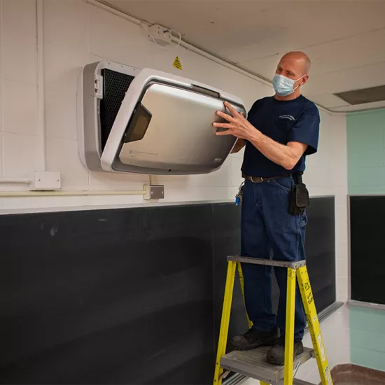 Man wearing blue shirt, jeans and blue paper face mask standing on step ladder in classroom installing an air purification unit on a brick wall above a chalkboard.