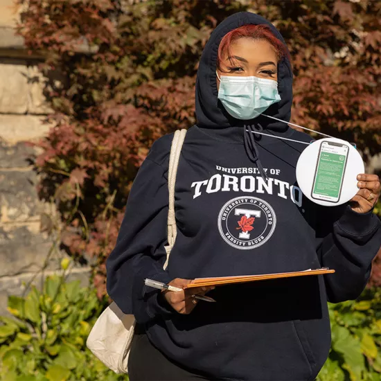 Woman wearing University of Toronto varsity hoodie and blue face mask holding up a lanyard with an image of the UCheck green screen on it.