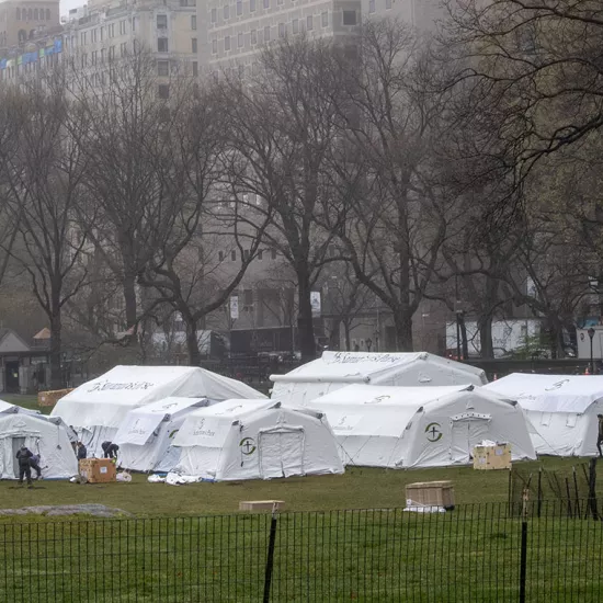 Large white tents in field with people working around them, apartment buildings in background