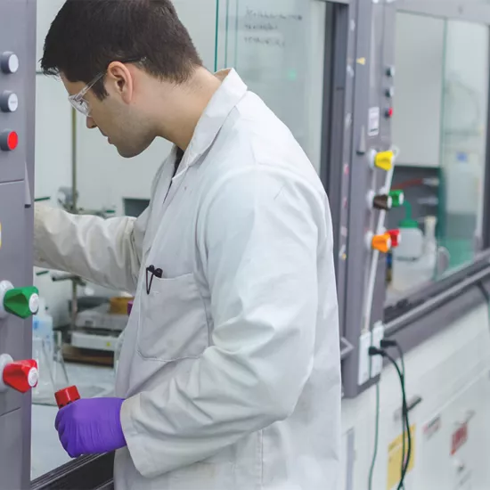 Aaron Cabral in lab coat, wearing safety glasses and purple latex-like gloves with a cylindar in his left hand. He is facing a bank of lab diagnostic equipment with colourful knobs.