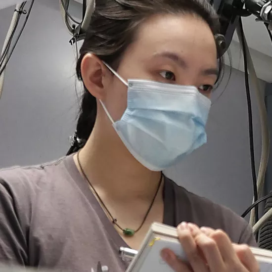 Student wearing blue surgical mask, jade necklace and brown t-shirt holding a notebook and pen, watching something off camera. Cables and unidentified equipment in the background.