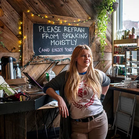 Sasha Steinberg standing by counter in restaurant, display of baked goods and four rows of shelves holding beverages behind her. Sign on counter says "Hot mulled cider" and chalkboard behind her on wall reads: Please refrain from speaking moistly (and enjoy your day).