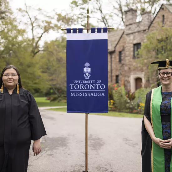 Kaeliana Smoke wearing a black robe and and Alexandra Gillespie wearing a black robe and green sash stand with a blue flag between them that reads University of Toronto Mississauga