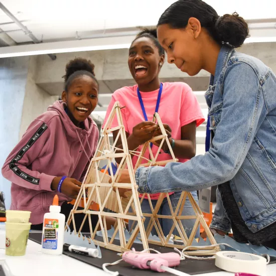 Three young students laugh as they build a pyramid from wooden popsicle sticks.