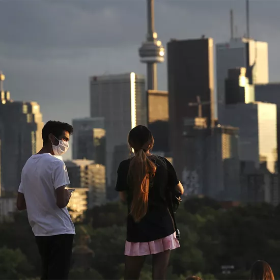 man and woman standing on hill overlooking at Toronto skyline. Man wearing mask, looking at woman who is, in turn, looking at skyline.