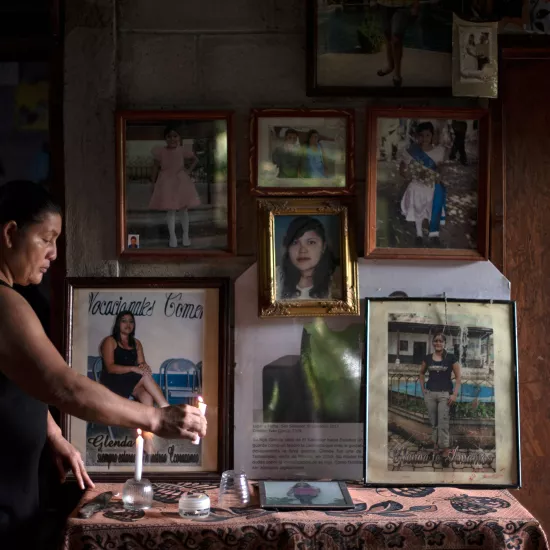Woman standing at table, lighting candle, various pictures of daughter seen hanging on wall behind table.