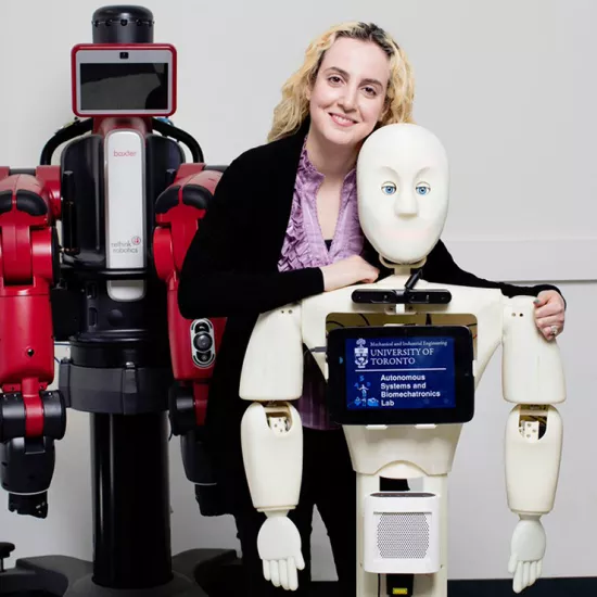 Woman standing next to two robots, one behind her is red, with a square screen for a face, the one in front of her is shorter, white, has a more human face with blue eyes and a screen on its chest that reads University of Toronto Autonomous Systems and Biomechatronics Lab