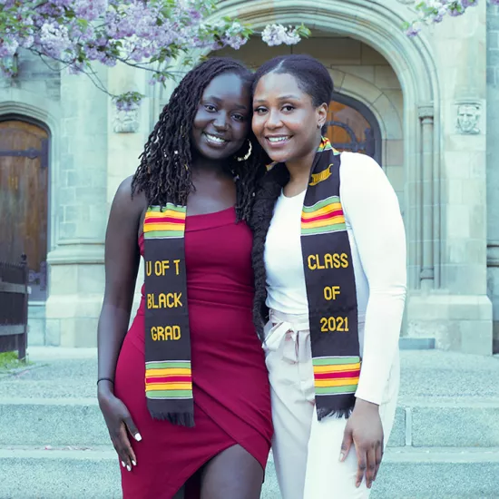 Two women standing in front of stone building wearing a black kente cloth that says U of T Black Grad Class of 2021