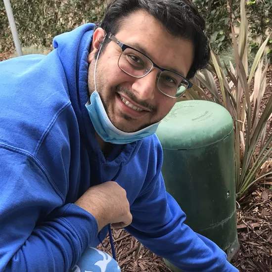 Jamsheed wearing a blue hoodie, glasses and blue face mask pulled down, smiling at camera while kneeling on a pathway next to greenspace with mulch on the ground.. 