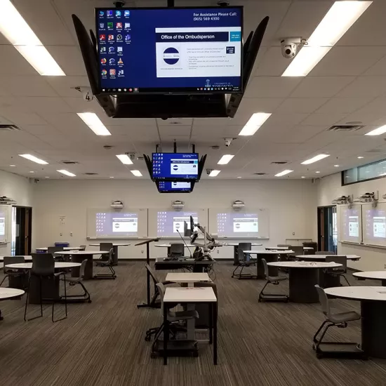 Room with round tables spread out, computer monitors hanging from ceiling, with image on monitors projected, separately, on three walls. Monitors show app icons on left side, slide in middle with title "Office of the Ombudsperson" and along top of screen it reads: For Assistance Phone 905-569-4300.Middle of room houses a small podium, two computer screens and a microphone