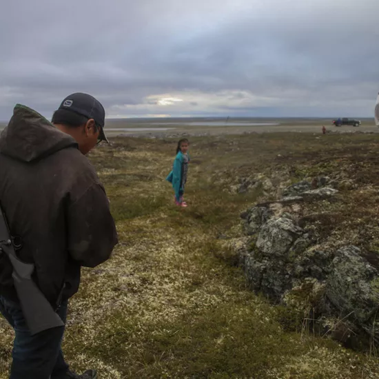 A man wearing a baseball hat with a rifle slung over his back walks along the tundra behind two girls, one stopping to look back, with a truck pickup truck in the background
