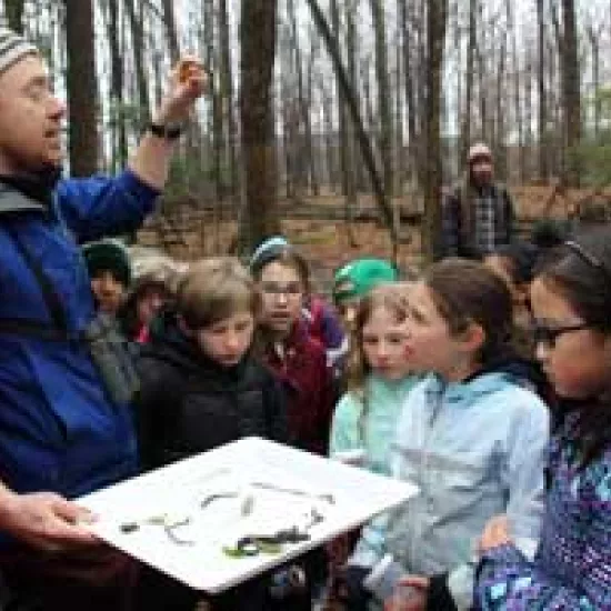 Associate professor of biology Marc Johnson stands in a forest setting surrounded by a group of attentive school children.