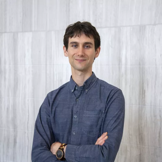 Michael Groechenig, wearing a dark blue button up shirt, standing in front of a concrete wall with his arms crossed.