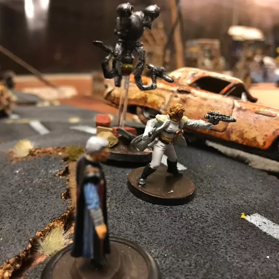 Photo of three miniature humanoid figures on an apocalyptic tabletop scene scattered with debris and overturned cars.