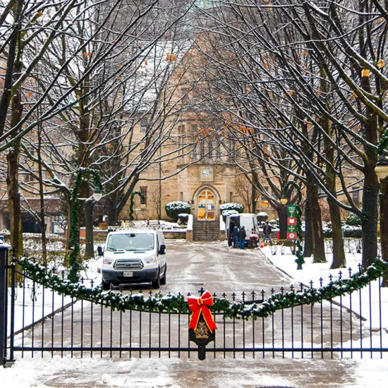 Closed iron gate with a garland and red bow on it, driveway leading up to old stone building on St. George campus