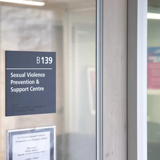 A glass door with a sign on it that reads: B139 Sexual Violence Prevention & Support Centre. Sign below reads: Tri campus sexual violence prevention and support. Office hours: 9am-5pm Monday, Tuesday, Thursday, Friday (rest of sign cut off from view)