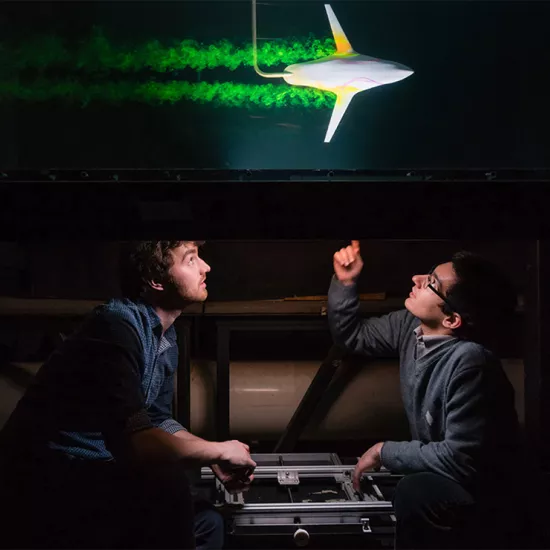 Two men sitting under a table, one pointing up. Top of table shows a small rocket-like device blasting buy with green smoke trailing behind